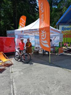 Powiększ zdjęcie: 1.	Zdjęcie przedstawia dwoje kobiety stojące pod namiotem obok  pomarańczowo zielonego plakat z napisem podziel się domem. Jedna kobieta w różowej bluzce trzyma rower.