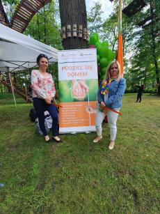 Powiększ zdjęcie: 2. Zdjęcie przedstawia dwie kobiety stojące obok plakatu z napisem podziel się domem, promującym rodzicielstwo zastępcze.