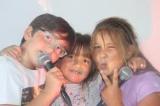 Powiększ zdjęcie: Trójka dzieci przytulona do siebie śpiewa do mikrofonu