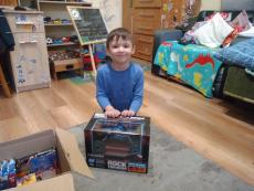 Powiększ zdjęcie: Zdjęcie przedstawia ciemnowłosego, radosnego chłopca siedzącego na podłodze, który z dumą prezentuje autko – prezent.