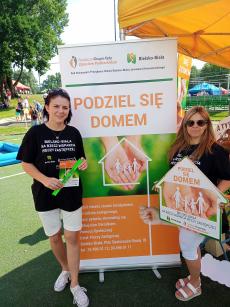 Powiększ zdjęcie: Zdjęcie przedstawia dwie kobiety stojące obok zielono pomarańczowego plakatu Podziel się domem, który promuje rodzicielstwo zastępcze