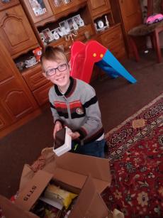 Powiększ zdjęcie: Zdjęcie przedstawia jasnowłosego, uśmiechniętego chłopca w okularach, który siedząc na podłodze rozpakowuje świąteczny prezent.