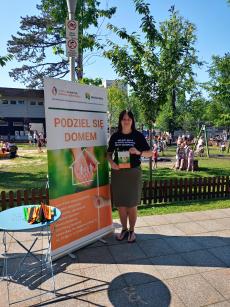 Powiększ zdjęcie: Zdjęcie przedstawia kobietę stojącą obok zielono pomarańczowego plakatu Podziel się domem, który promuje rodzicielstwo zastępcze.