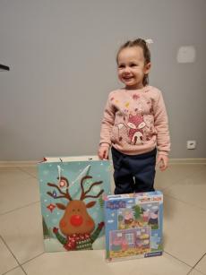 Powiększ zdjęcie: Zdjęcie przedstawia uśmiechniętą dziewczynkę stojącą obok dwóch prezentów