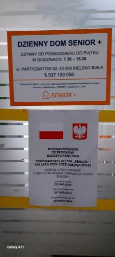 Powiększ zdjęcie: Drzwi wejściowe do Dziennego Domu Senior+ przy ul. Partyzantów 62 w Bielsku-Białej. Na górze plakat z białym tłem i pomarańczową ramką. Granatowymi literami jest napisane: Dzienny Dom Senior+ czynny od poniedziałku do piątku w godzinach 7.30-15.30, ul. Partyzantów 62, 43-300 Bielsko-Biała, telefon 537 193 056. Dzienny Dom Senior+ korzysta z dofinansowania ze środków otrzymanych w ramach Programu Wieloletniego „Senior+” na lata 2015-2020. Na dole logo Programu Senior+ czyli pomarańczowy domek z sercem. Poniżej Plakat Informacyjny Programu SENIOR+, który przedstawia u góry flagę i godło Polski, poniżej napis: Dofinansowano ze środków budżetu państwa. Program Wieloletni „Senior+” na lata 2021-2025 edycja 2024. Moduł II – Zapewnienie funkcjonowania Dziennego Domu Senior+. Na dole plakatu napis: dofinansowanie 123 840,00 zł całkowita wartość 267 840,00 zł data podpisania umowy czerwiec 2024.