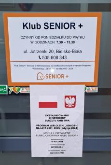 Powiększ zdjęcie: Drzwi wejściowe do Klubu Senior+ przy ul. Jutrzenki 20 w Bielsku-Białej. Na górze drzwi plakat z białym tłem i pomarańczową ramką. Granatowymi literami napisano: Klub Senior+ czynny od poniedziałku do piątku w godzinach 7.30-15.30, ul. Jutrzenki 20, 43-300 Bielsko-Biała, telefon 535 608 343. Klub Senior+ korzysta z dofinansowania ze środków otrzymanych w ramach Programu Wieloletniego „Senior+” na lata 2015-2020. Na dole logo Programu Senior+ czyli pomarańczowy domek z sercem. Poniżej na drzwiach Plakat Informacyjny Programu SENIOR+, który przedstawia u góry flagę i godło Polski, poniżej napis: Dofinansowano ze środków budżetu państwa. Program Wieloletni „Senior+” na lata 2021-2025 edycja 2024. Moduł II – Zapewnienie funkcjonowania Klubu Senior+. Na dole napis: dofinansowanie 61 920,00 zł całkowita wartość 141 920,00 zł data podpisania umowy czerwiec 2024.