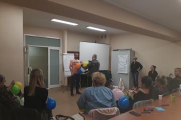 Zdjęcie przedstawia osoby uczestniczące w szkoleniu dla kandydatów na rodziny zastępcze niezawodowe.  Na pierwszym planie widać uczestników szkolenia siedzących w kręgu. Natomiast w głębi stoją trzy osoby, które wykonują ćwiczenie zalecone przez trenera.